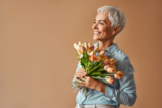 Portrait of a beautiful short-haired gray-haired confident charming mature woman holding a bouquet of orange tulips while standing on a beige background and looking into space.