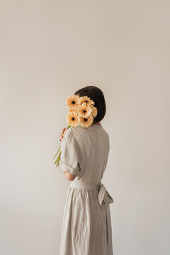 Beautiful young woman in neutral sandy linen dress holding gerber flowers bouquet over white wall. Aesthetic fashion background
