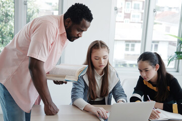 Young African teacher or tutor is holding book and teaching for knowledge about online research with internet on laptop for Caucasian student girls in classroom. Education and Technology Concept
