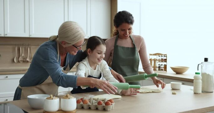 Cheerful mature grandma teaching cute granddaughter kid and daughter woman to bake, helping girl to roll dough on kitchen table, speaking, smiling, laughing, having fun with family
