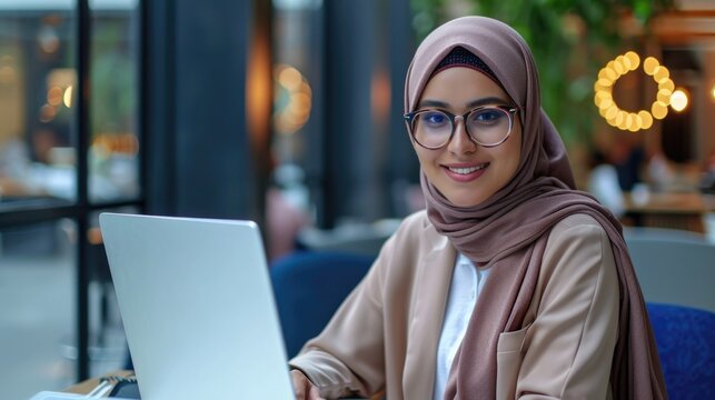Young and successful arabian business woman in hijab working with laptop inside modern office building, muslim woman in glasses smiling and happy with work achievement.
