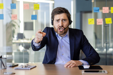 Webcam view, angry man with headset phone yelling at webcam, computer screen, businessman arguing with colleagues and partners inside office at workplace, video call online meeting with employees.