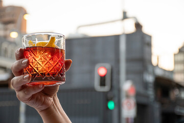 female hand raising with glass of red negroni drink with orange slices in blurred city traffic light