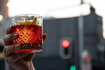 female hand raising with glass of red negroni drink with orange slices in blurred traffic light