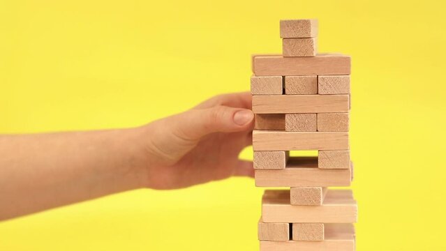 A woman plays Jenga on a yellow background. The concept of family vacation, holidays, entertainment. A woman's hand takes out a block from a wooden toy tower. A game