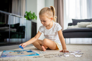 Fototapeta premium Little blonde girl sits at home on the carpet and collects puzzles