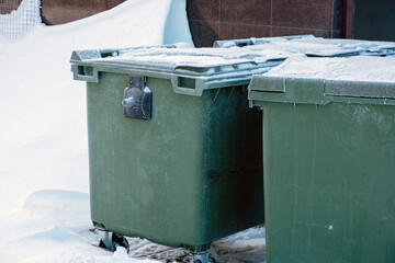 Green plastic dumpsters in a snowdrift on a winter street are covered with frost