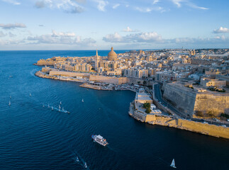 Fototapeta na wymiar Valletta old town with cathedral at golden sunset. Aerial view on historic city with sailing boats returning into harbor. Malta at the end of summer season.