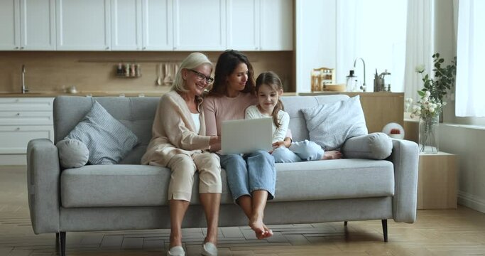 Happy senior grandma, mom and cute little girl using laptop on comfortable couch together, enjoying modern technology, online communication, shopping on Internet, media service for entertainment