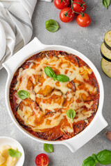 Eggplant casserole with cheese and tomato sauce in a white baking dish on a gray concrete background with ingredients for cooking. Vegetarian healthy food.