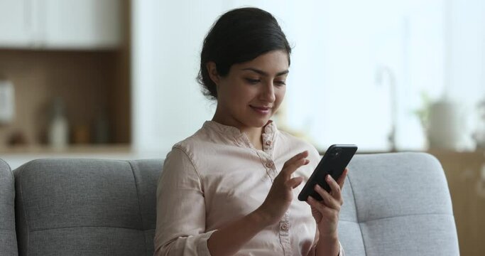 Positive pretty Indian woman using online app on smartphone for communication, sitting on home couch, typing message, chatting, getting cheerful, smiling, turning look away, thinking