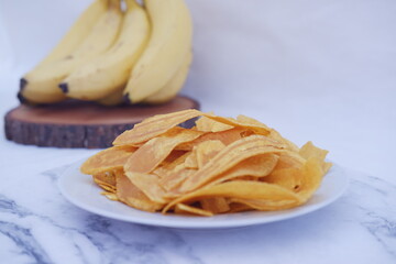 Crispy banana chips on a white plate with fresh bananas.