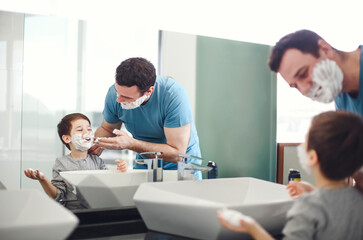 Fototapeta premium Young father and little son applying shaving foam on face in bathroom at the morning together near the mirror