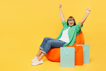 Full body elderly woman 50s year old in green shirt casual clothes sit in bag chair hold shopping package bags do winner gesture isolated on plain yellow background Black Friday sale buy day concept