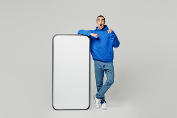 Full body young middle eastern man in blue hoody casual clothes big huge blank screen mobile cell phone with workspace copy space area point index finger up isolated on plain solid white background
