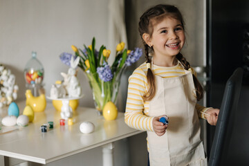 Process of little girl painting eggs on kitchen. Spring holiday. Easter decoration at home. Side...