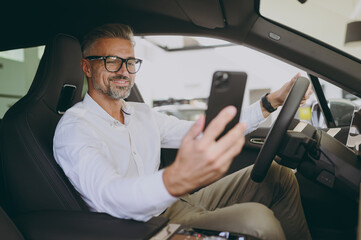 Adult happy man customer male buyer client wear shirt do selfie on mobile cell phone drive car choose auto want buy new automobile in showroom vehicle salon dealership store motor show. Sales concept. - obrazy, fototapety, plakaty