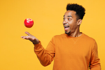 Young shocked man wear orange sweatshirt casual clothes hold in hand toss up red apple isolated on plain yellow background studio portrait. Proper nutrition healthy fast food unhealthy choice concept.