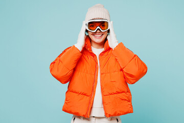 Young smiling happy woman wear warm padded windbreaker jacket hat ski put hands on goggles mask looking camera travel rest spend weekend winter season in mountains isolated on plain blue background. © ViDi Studio