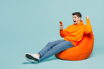 Full body fun young man he wears orange hoody casual clothes sit in bag chair hold use mobile cell...