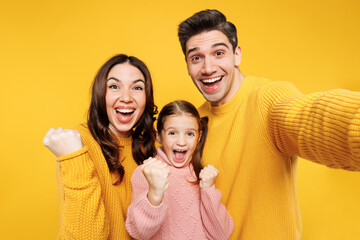 Fototapeta premium Close up winner fun young parents mom dad with child kid girl 7-8 years old wear pink sweater casual clothes do selfie shot on mobile cell phone isolated on plain yellow background Family day concept