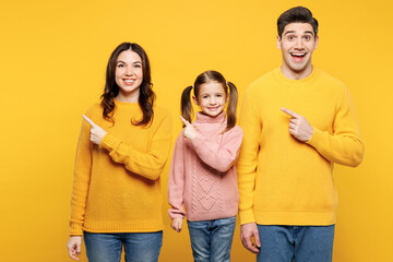 Fototapeta premium Young happy parents mom dad with child kid girl 7-8 years old wear pink knitted sweater casual clothes point index finger aside on empty area isolated on plain yellow background. Family day concept.