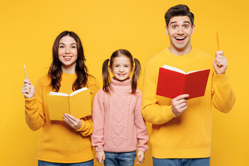 Young happy cool parents mom dad with child kid girl 7-8 years old wear pink sweater casual clothes read book point finger pen up with new idea isolated on plain yellow background. Family day concept.