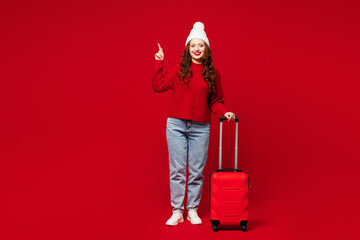 Traveler woman in sweater hat casual clothes hold bag point finger aside isolated on plain red...