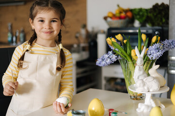 Cute four year girl in beige apron sitting on kitchen table and painting easter eggs