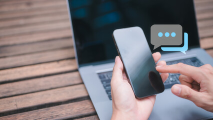 hands using smartphone with pop up chatting icon and blurred computer on outdoor table background in concept of social media internet network online marketing