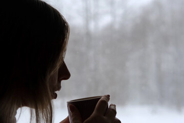 Long hair woman standing among the window and drinking hot tea from mug. Winter landscape. Lady...