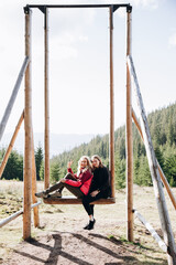 two women travel through the mountains. go on a hike with friends. girlfriends are sitting on a large wooden swing in the middle of the mountains