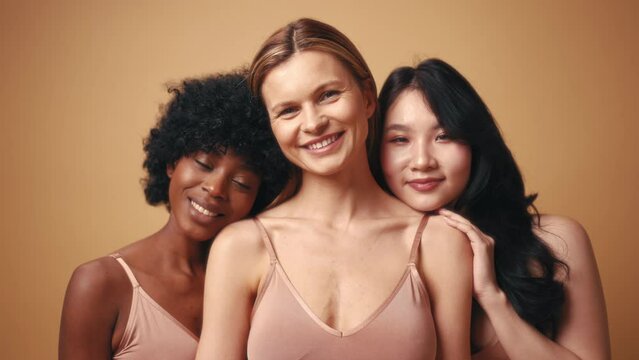 Multi-ethnic multiracial young women in beige lingerie isolated on a beige background. African-American, Caucasian and Asian women posing in the studio. Concept of face body skin care, cosmetology.