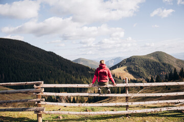 a woman in a pink ski jacket sits on a wooden fence overlooking the mountains. travel in the...