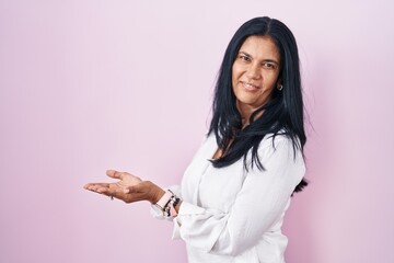 Mature hispanic woman standing over pink background pointing aside with hands open palms showing...