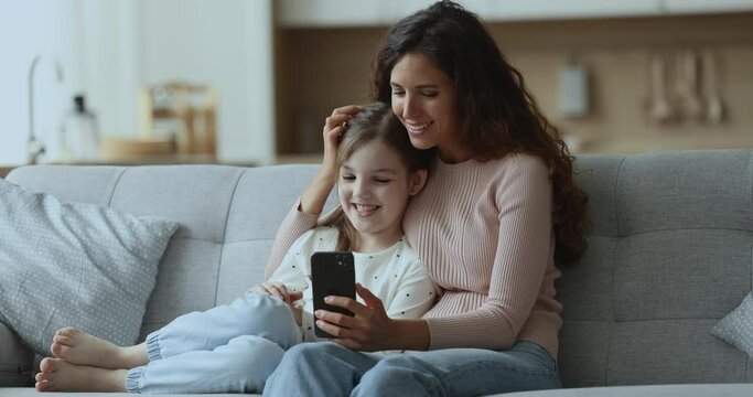 Cheerful beautiful Latin mom and cute daughter girl watching online video content on smartphone, hugging on comfortable home couch, using mobile phone for online entertainment
