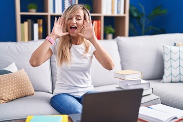 Young blonde woman studying using computer laptop at home smiling cheerful playing peek a boo with...