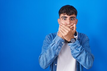 Young hispanic man standing over blue background shocked covering mouth with hands for mistake. secret concept.
