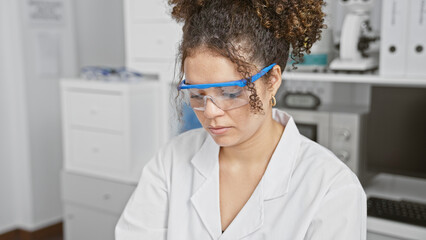A young hispanic woman with curly hair works attentively in a laboratory setting, wearing safety goggles and a lab coat.