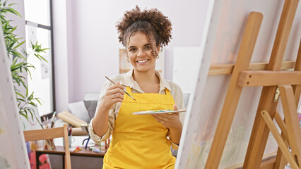 A smiling young hispanic woman with curly hair paints on a canvas in a bright art studio.