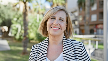 A smiling caucasian middle-aged woman in a striped jacket outdoors in a sunny park.