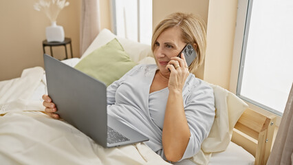 Mature woman multitasking with a laptop and phone in a cozy bedroom setting