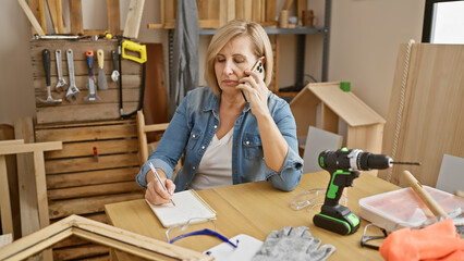 A middle-aged woman multitasks in a woodworking workshop, talking on the phone while taking notes.