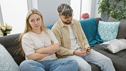 A displeased woman and a distracted man sit together but emotionally apart on a couch in a modern...