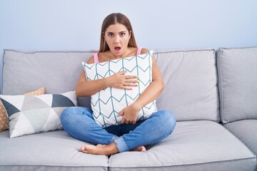 Young blonde woman hugging pillow sitting on the sofa in shock face, looking skeptical and sarcastic, surprised with open mouth