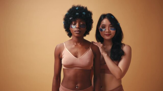 African-American and Asian young women using eye patches in lingerie posing on beige background. Cosmetology and spa treatments for face and body.
