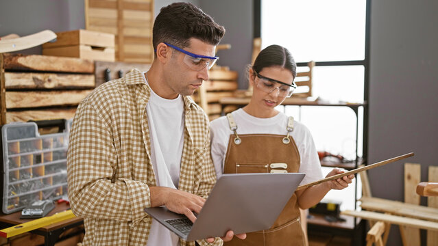 Man and woman in workshop looking at laptop reviewing designs amidst woodworking tools.