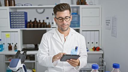 Naklejka na ściany i meble Handsome young hispanic man, confident smiling scientist, comfortably working with touchpad at his high-tech lab, advancing in the world of science and medicine