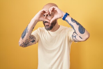 Hispanic man with tattoos standing over yellow background doing heart shape with hand and fingers...