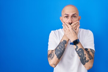 Hispanic man with tattoos standing over blue background shocked covering mouth with hands for...